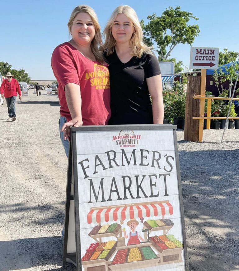 Two smiling women standing behind the farmer's market sign.